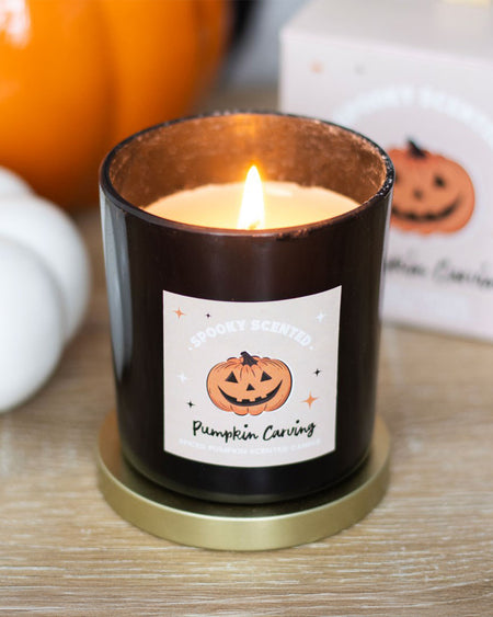 A close-up of the lit Pumpkin Carving candle, with its rich spiced pumpkin fragrance and black glass jar, sitting on a golden coaster with autumnal decor in the background.