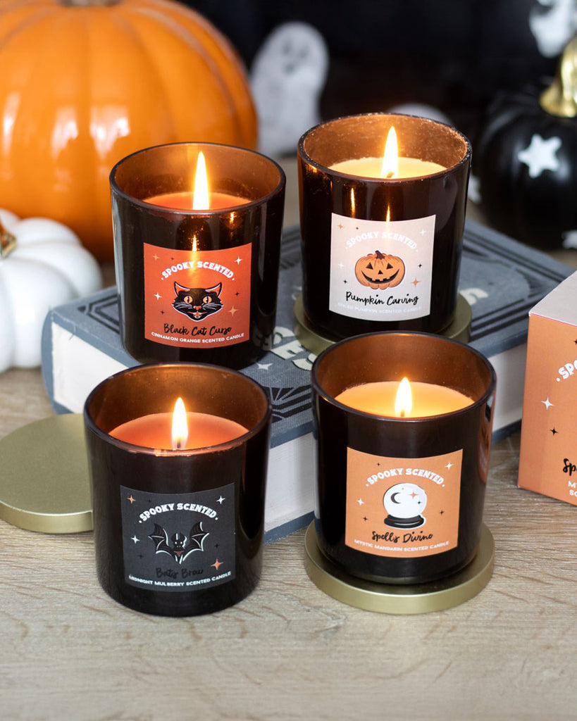 A cosy Halloween-themed setup featuring four spooky scented candles in black glass jars, each with a unique label. The Pumpkin Carving candle, with a jack-o'-lantern label, emits a warm glow, surrounded by pumpkins and eerie decorations.
