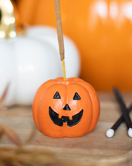 Pumpkin-shaped incense holder with a happy jack-o'-lantern face holding a burning incense stick, styled on a wooden tray with autumn-themed props. A fun and spooky Halloween home accent from Rebel Romance.