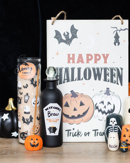 Halloween-themed flat lay featuring spooky decorations including a black candle, "Witches Brew" bottle, skeleton nesting dolls, and an orange jack-o'-lantern incense stick holder with a burning incense stick. Perfect gothic autumn home décor from Rebel Romance.