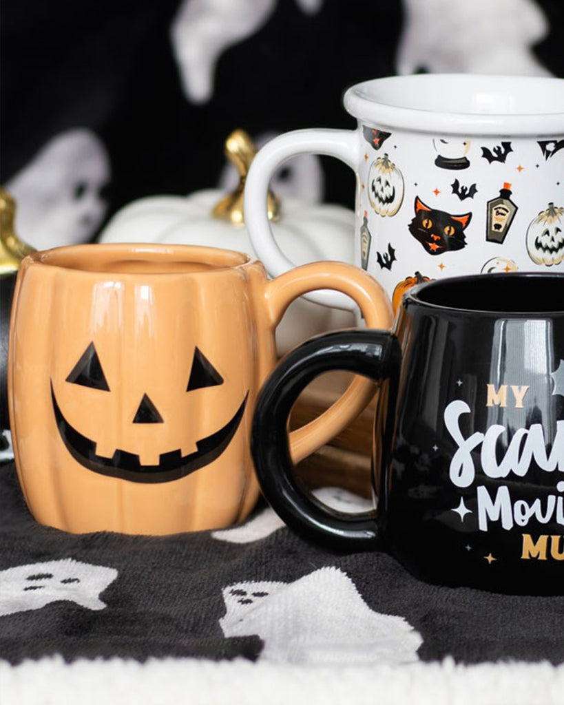 Three Halloween mugs including a pumpkin-shaped mug, a black "scary movie" mug and a white spooky print mug, styled with black and white ghost blanket for autumn aesthetic.
