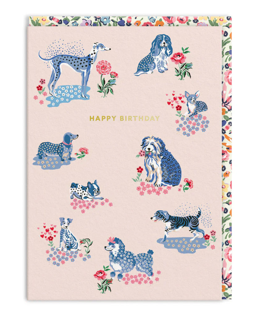 A Cath Kidston A6 birthday card featuring a pastel pink background with illustrated blue-toned dogs in floral patches, surrounded by pink, red, and blue flowers. Includes a gold foil ‘Happy Birthday’ message in the centre and a floral patterned envelope. From Rebel Romance, a UK brand celebrating quirky, alternative aesthetics.