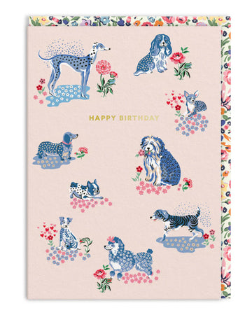 A Cath Kidston A6 birthday card featuring a pastel pink background with illustrated blue-toned dogs in floral patches, surrounded by pink, red, and blue flowers. Includes a gold foil ‘Happy Birthday’ message in the centre and a floral patterned envelope. From Rebel Romance, a UK brand celebrating quirky, alternative aesthetics.