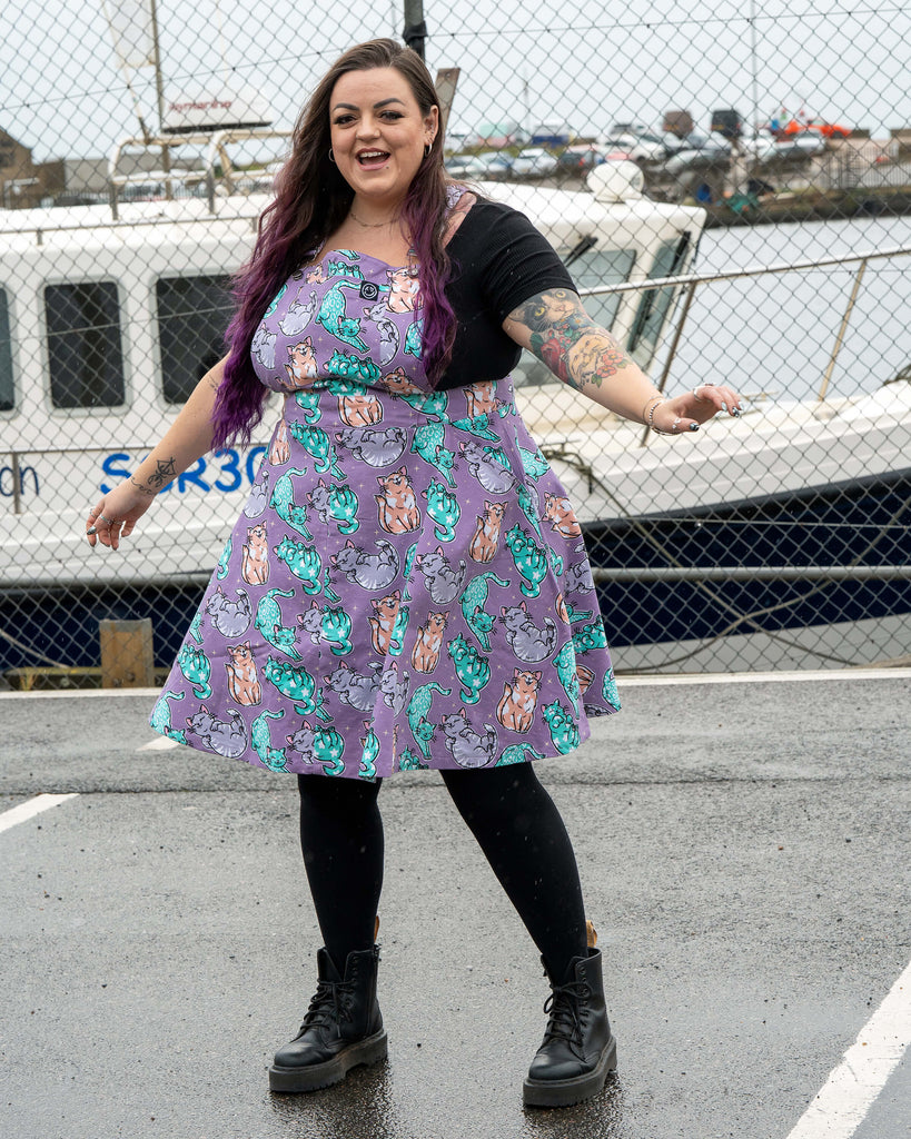 Luisa smiles while wearing the Rebel Romance Purrfect Cats Flared Pinafore. The lavender skater style dress twirls as she moves, showing the pastel illustrated cats all over the fabric. She styles it with a black top, tights and chunky boots, standing near boats and a metal fence.