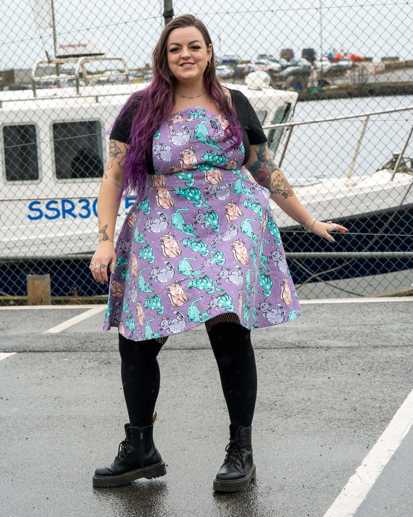 Luisa poses in front of a harbour scene wearing the Rebel Romance Purrfect Cats flared pinafore. The lilac cat print skirt spreads as she holds it out. She smiles happily with purple hair, tattoos and black boots completing the look.