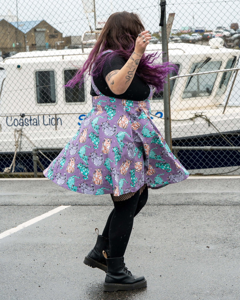 Side view of Luisa spinning in the Rebel Romance Purrfect Cats Flared Pinafore. The flare of the skirt shows the full pastel cat pattern in mint, peach, grey and teal. She wears black tights and black boots, with the harbour boats and fencing in the background.