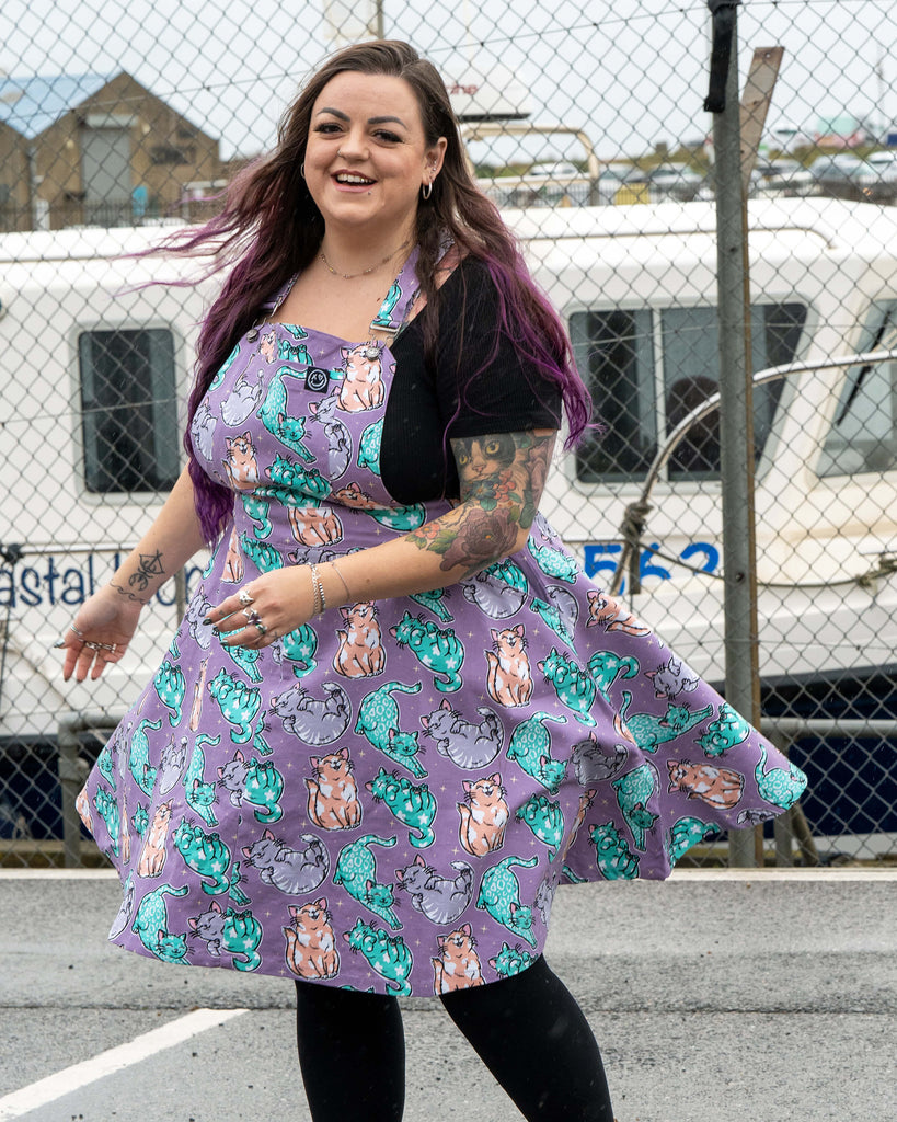 Luisa twirls in the Rebel Romance Purrfect Cats Flared Pinafore, the skater skirt fanning out to show the pastel cat print clearly. She wears a black top and leggings with black boots, standing on a wet seaside car park beside a boatyard.