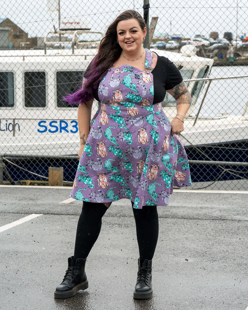 Luisa stands smiling in the Rebel Romance Purrfect Cats Flared Pinafore. The soft lavender fabric is covered in illustrated pastel cats. She is wearing black boots, tights and a black top, photographed in front of a white boat and metal fence.