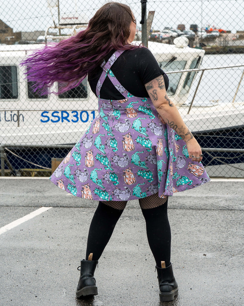 Luisa poses in the Rebel Romance Purrfect Cats flared pinafore with a lilac cat print. She twirls in a harbour car park, smiling with purple hair flowing. The pinafore has a fitted bib, flared skirt and adjustable straps. She wears black leggings, a black top and chunky boots.