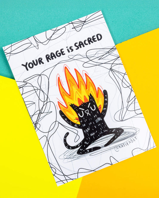 A postcard of an angry black cat on fire on a white background with back squiggles it has text above saying 'Your rage is sacred'. Illustrated by Katie Abey.
