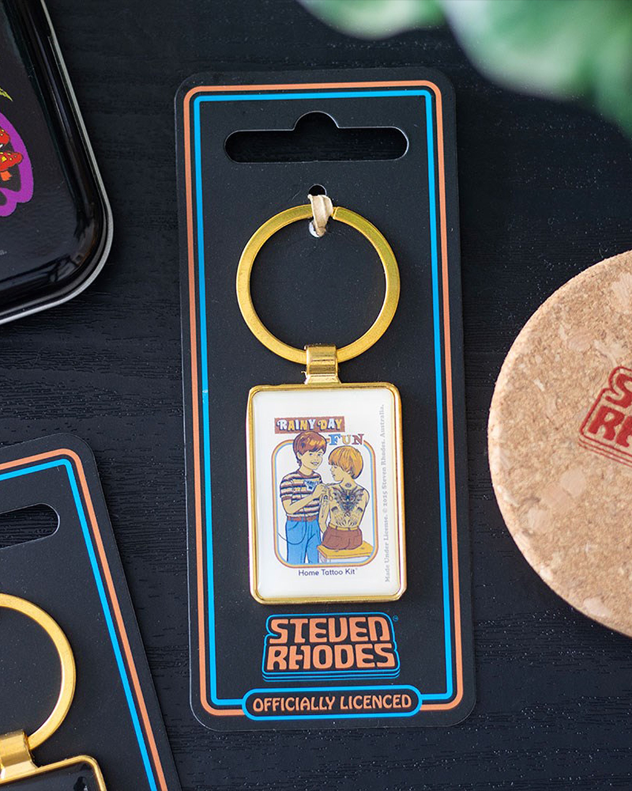 Rainy Day Fun Keyring by Steven Rhodes on backing card packaging, officially licensed. Sold by Rebel Romance, a UK alternative fashion and lifestyle store. The enamel gold keyring features retro tattoo art, making it a unique gift idea for quirky and rebellious personalities.