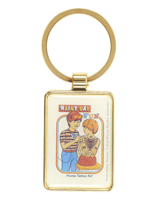 Front view of the Steven Rhodes Rainy Day Fun Keyring from Rebel Romance. The enamel keyring has a shiny gold finish and features vintage-inspired tattoo artwork with the text “Home Tattoo Kit.” A quirky, alternative gift for millennials who love retro humour and unique accessories.