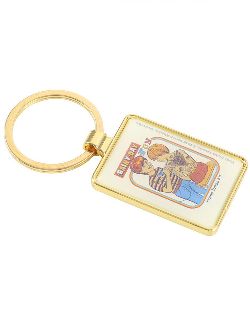 Rainy Day Fun Keyring by Steven Rhodes displayed on its side. This enamel gold keyring from Rebel Romance shows a retro illustration of children tattooing at home. A funny, alternative accessory that makes a bold statement, perfect for keys, bags, or as a unique retro gift.