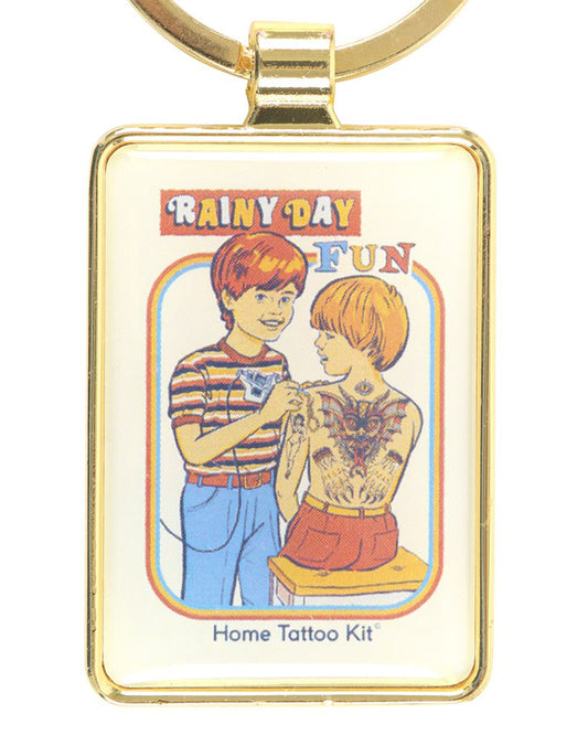 Close-up of the Rainy Day Fun Keyring by Steven Rhodes, sold by Rebel Romance. The enamel keyring has a gold frame and features retro-style art of two children with a “Home Tattoo Kit.” Quirky and alternative, this officially licensed keyring is a playful gift for tattoo lovers and fans of dark humour.