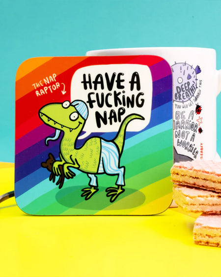 The Nap Raptor coaster propped in front of a confidence themed mug by Katie Abey. The green dinosaur character with sleepy vibes is shown against a bold rainbow-striped background.