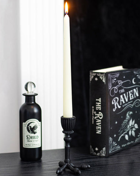 Black taper candle holder shaped like a raven’s claw, made from resin with a matte finish. Shown on a gothic-style black surface beside The Raven book and a potion bottle, holding a tall white taper candle. Perfect for Halloween or gothic home decor.
