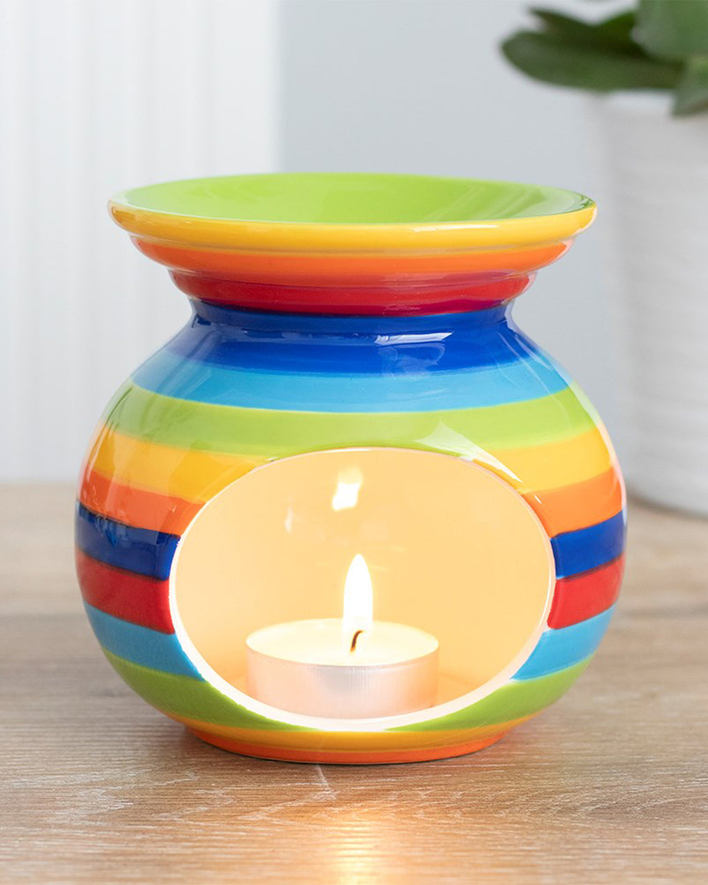 A vibrant Rainbow Stripe Oil Burner by Rebel Romance sits on a wooden surface with a lit tealight inside. The glossy ceramic burner features bold, colourful stripes and a lime green bowl for wax melts or oils.