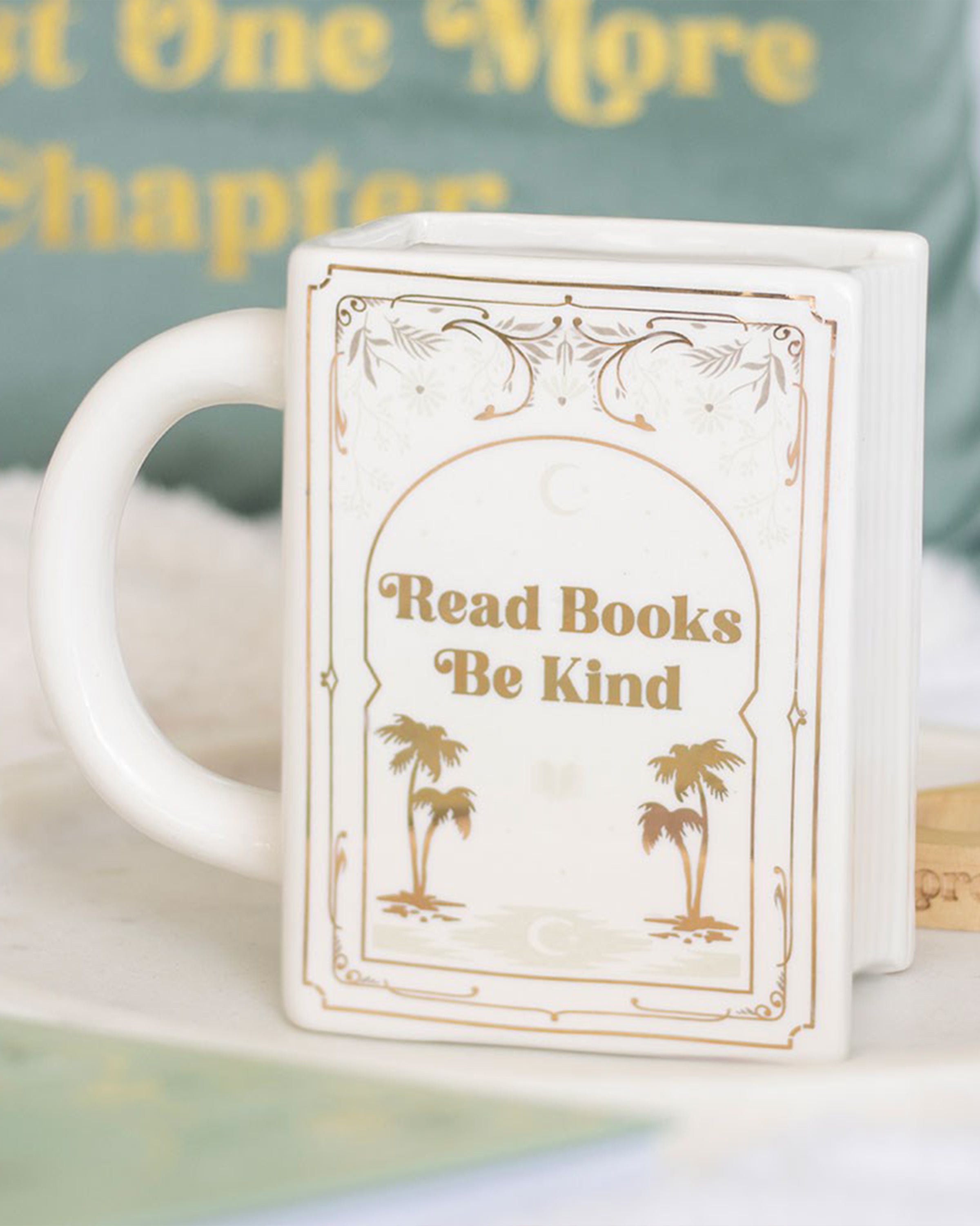 Styled image of the Rebel Romance Read Books Be Kind ceramic mug placed on a white surface with a pastel book-themed cushion in the background. A thoughtful and whimsical homeware gift for book lovers and fans of cosy aesthetics.