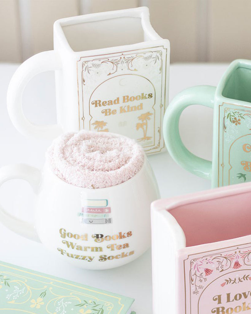 A white ceramic book-shaped mug from Rebel Romance with the phrase “Read Books Be Kind” in gold lettering. Styled alongside pastel mugs and a cosy sock-filled round mug, this homeware gift is perfect for book lovers, tea drinkers, and fans of unique mugs. A quirky literary-inspired design from a small UK alternative brand.