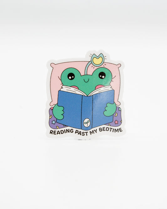 Adorable die-cut vinyl sticker of a green frog tucked in bed with a crown, reading a blue book. Surrounded by a pink pillow and starry pyjamas, with the caption “Reading Past My Bedtime.” A cosy design perfect for bookworms and frog lovers. Created by Rebel Romance, a small UK brand celebrating alternative fashion, slow style, and quirky, millennial-friendly gifts with charm.
