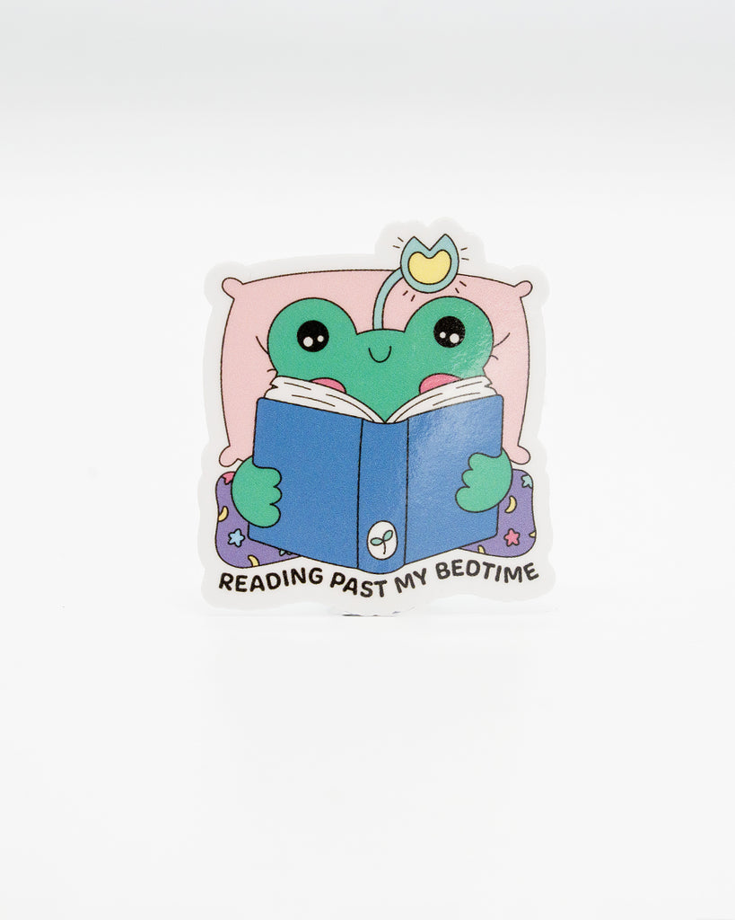 Adorable die-cut vinyl sticker of a green frog tucked in bed with a crown, reading a blue book. Surrounded by a pink pillow and starry pyjamas, with the caption “Reading Past My Bedtime.” A cosy design perfect for bookworms and frog lovers. Created by Rebel Romance, a small UK brand celebrating alternative fashion, slow style, and quirky, millennial-friendly gifts with charm.