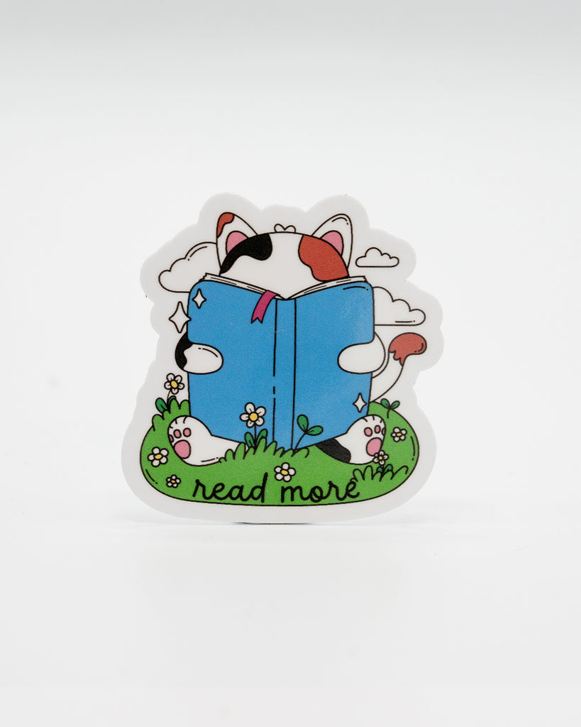 Adorable die-cut vinyl sticker of a calico cat reading a big blue book while lying in a grassy field with daisies. Surrounded by clouds and the phrase “read more” at the bottom. A perfect design for book lovers and cat fans alike. Created by Rebel Romance, a small UK brand known for alternative style, slow fashion, and cute, millennial-friendly bookish gifts and accessories.