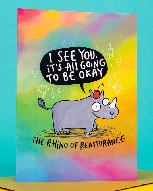 A colourful A6 greeting card by Rebel Romance featuring a cute cartoon rhino wearing a red fez, with the words “I see you. It’s all going to be okay” in a speech bubble. Below the rhino, it says “The Rhino of Reassurance”. The background is a cheerful rainbow gradient with sparkles. Designed by Katie Abey and perfect for sending positivity, comfort, and humour. Ideal for alternative millennial shoppers looking for quirky, uplifting cards.