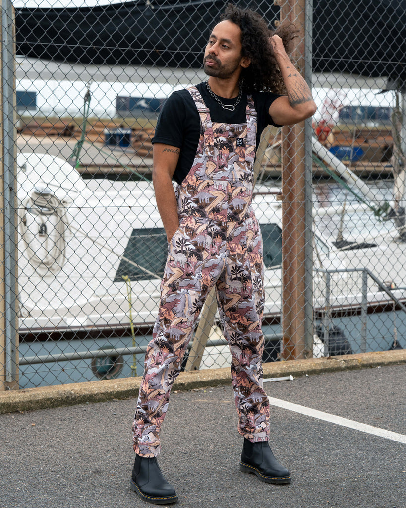Richard posing outdoors by a chain-link fence in Run & Fly Dinoverse Stretch Twill Dungarees, showing front bib pocket and bold retro dinosaur print.