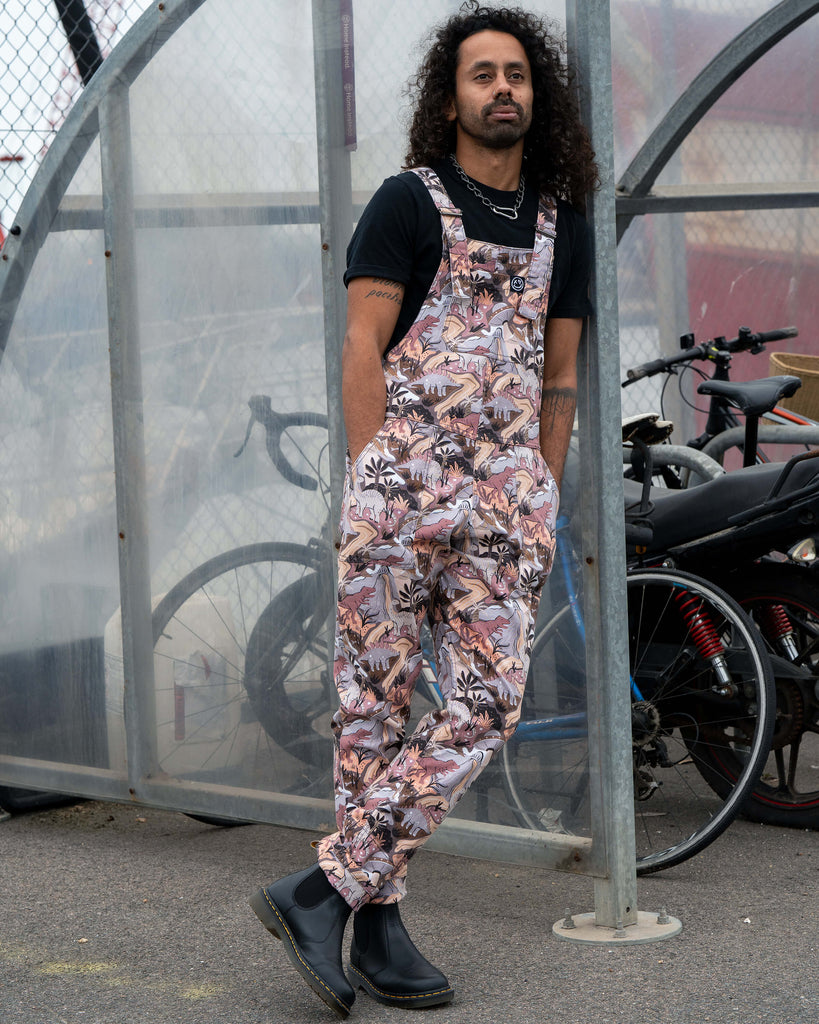 Richard wearing Rebel Romance Dinoverse Stretch Twill Dungarees with hands in pockets, leaning casually by a bike rack, showing the playful dinosaur print.