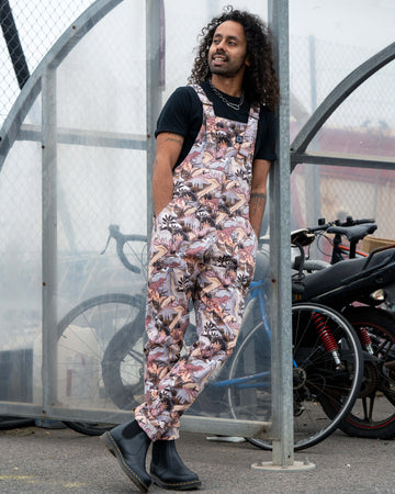 Richard wearing Rebel Romance Dinoverse Stretch Twill Dungarees with hands in pockets, leaning casually by a bike rack, showing the playful dinosaur print.