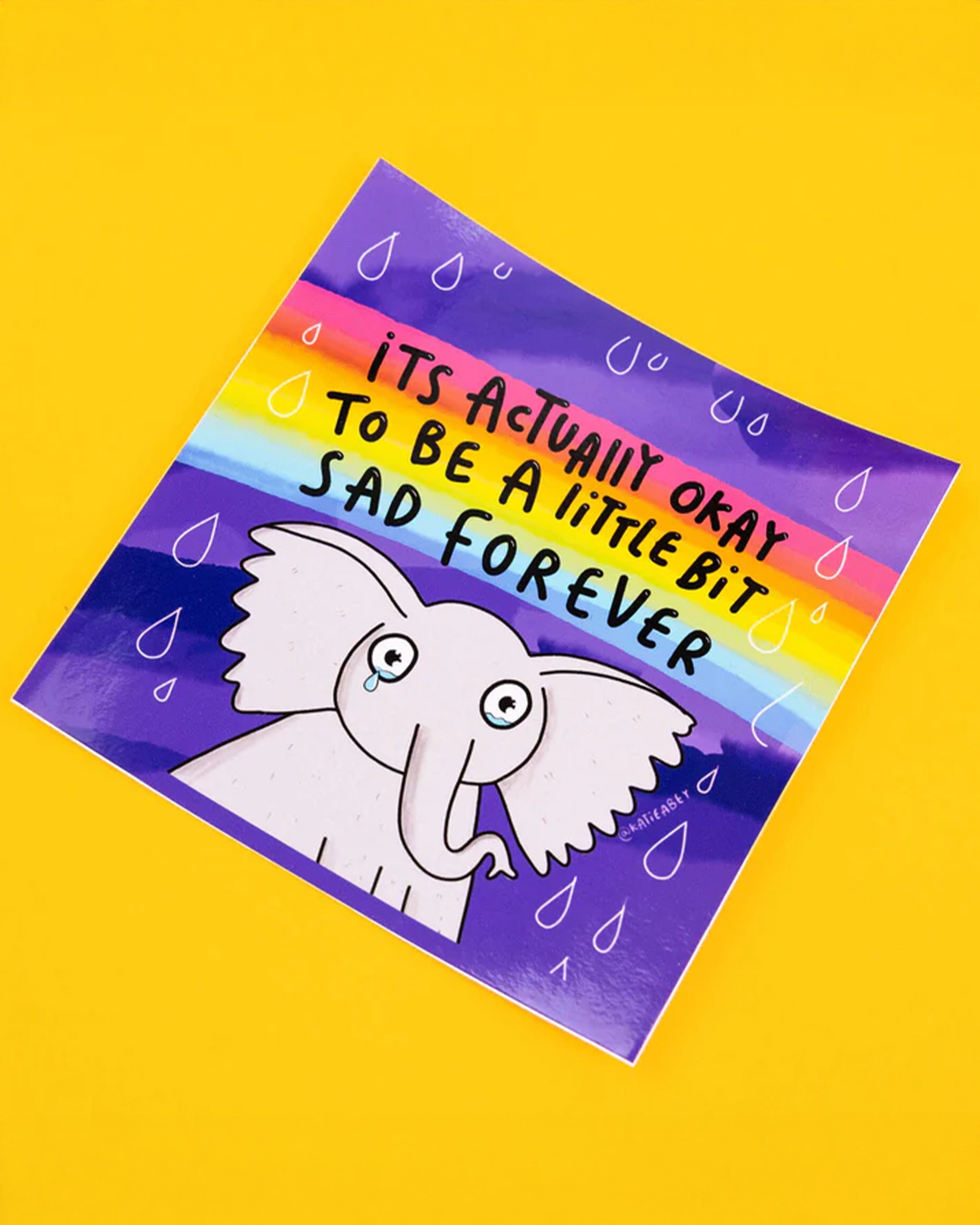 Katie Abey vinyl sticker stocked at Rebel Romance of a sad looking cartoon elephant with a tear, set against a purple background with a rainbow above. The sticker reads 'It’s Actually Okay to Be a Little Bit Sad Forever' in comforting black letters, with small raindrop details around the text.