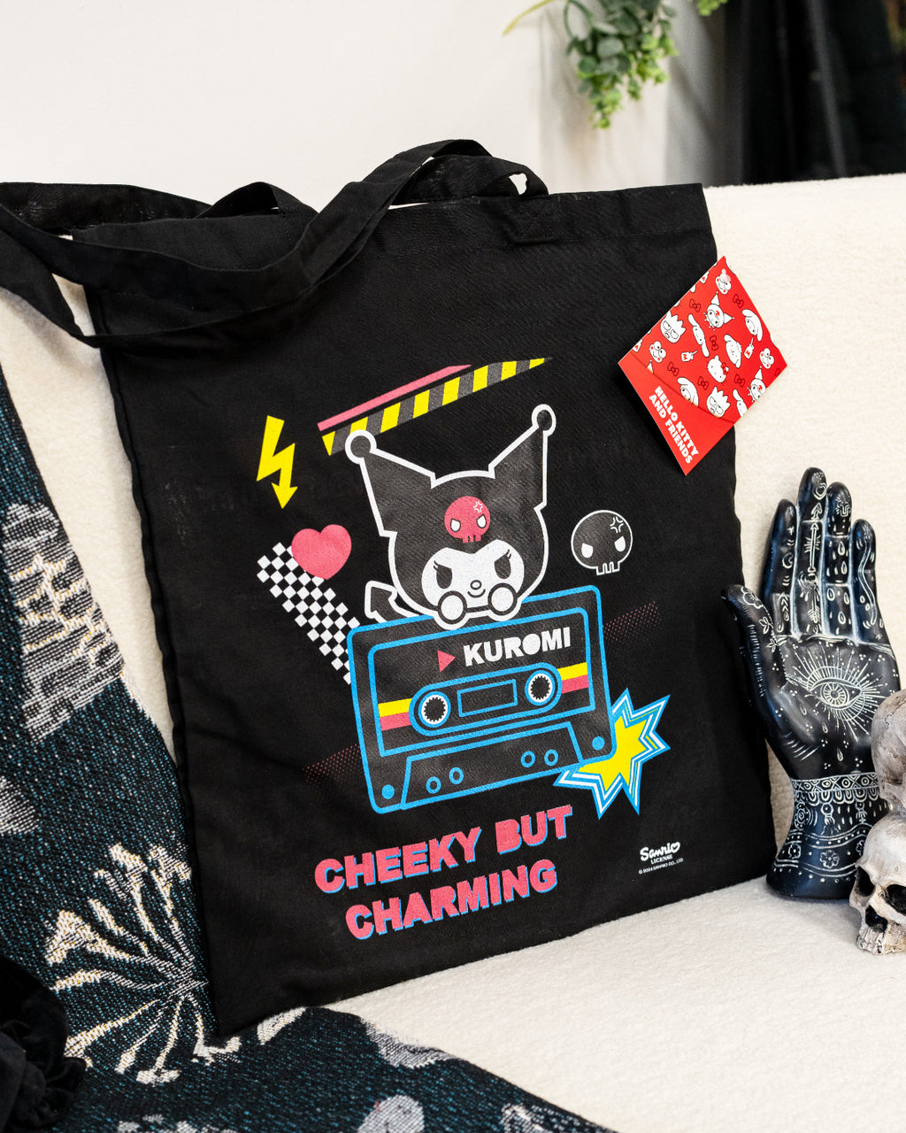 Kuromi Cheeky But Charming Tote Bag displayed in a cosy setting, showcasing a vibrant design with Kuromi, a cassette tape motif, and colourful details. A standout piece from Rebel Romance, a UK-based brand embracing alternative style and retro-inspired products. Ideal for bold, playful fashion lovers.