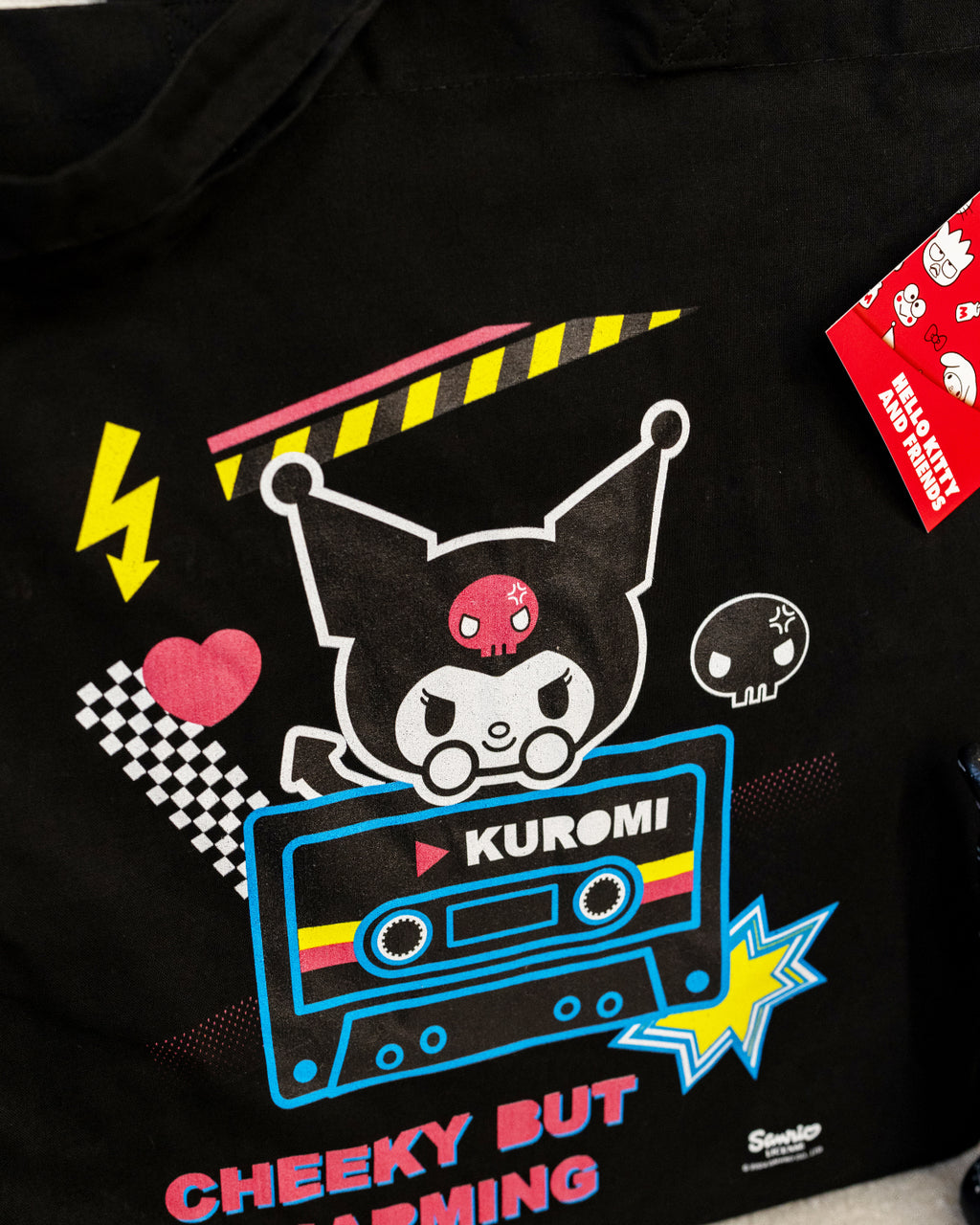 Close-up of the Kuromi Cheeky But Charming Tote Bag featuring the mischievous Sanrio character Kuromi atop a retro cassette tape design, surrounded by bright accents, a heart, skulls, and bold text. Sold by Rebel Romance, a small UK brand celebrating alternative fashion and retro aesthetics. Perfect for adding quirky flair to your daily essentials.
