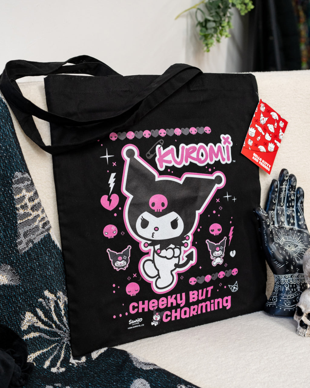 The Kuromi Cheeky Black Tote Bag displayed on a cosy setting, featuring Kuromi’s playful design surrounded by pink skulls, hearts, and bold text. A standout piece from Rebel Romance’s alternative collection, perfect for adding a quirky flair to your outfits.