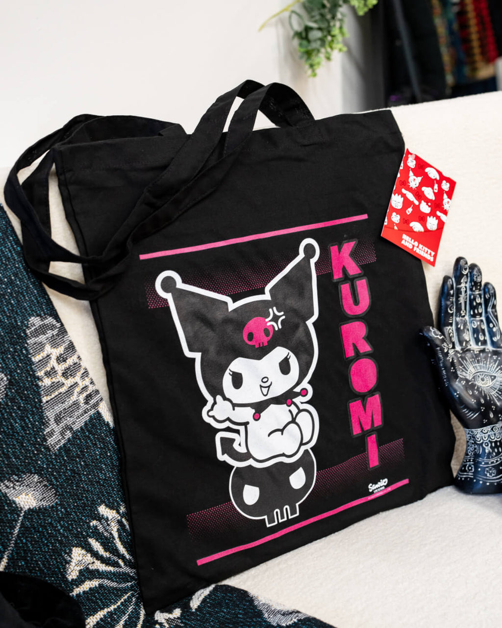 A black tote bag featuring Kuromi in her classic mischievous pose on a skull, with bold pink accents and the text “Kuromi” vertically on the side. The bag is stylish, edgy, and perfect for fans of the Sanrio character.