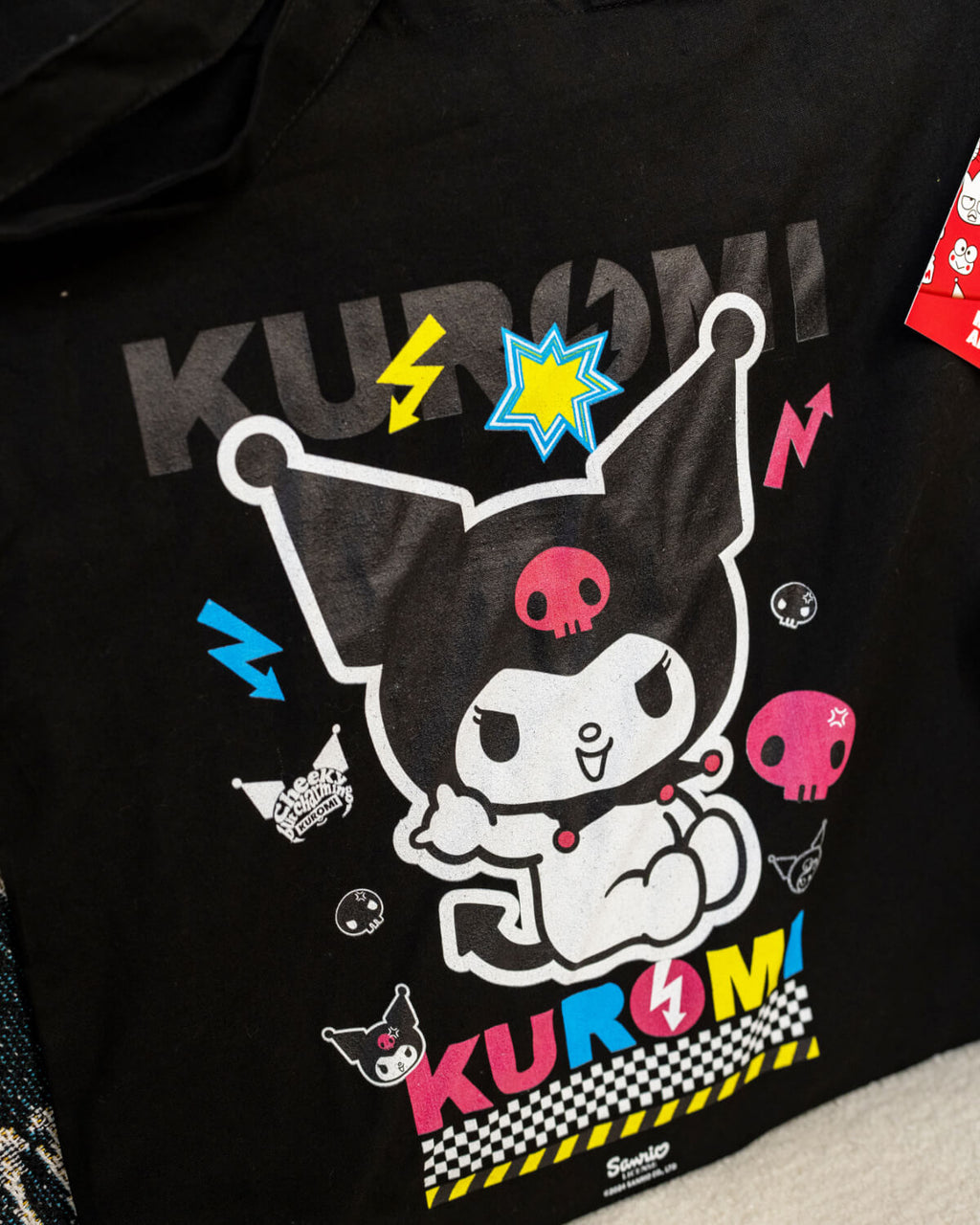 A black tote bag featuring Sanrio’s Kuromi in a colourful design with neon pink, blue, and yellow accents. The bag showcases Kuromi surrounded by lightning bolts, skulls, and fun punk-style details. Perfect for anime fans and alternative fashion lovers.