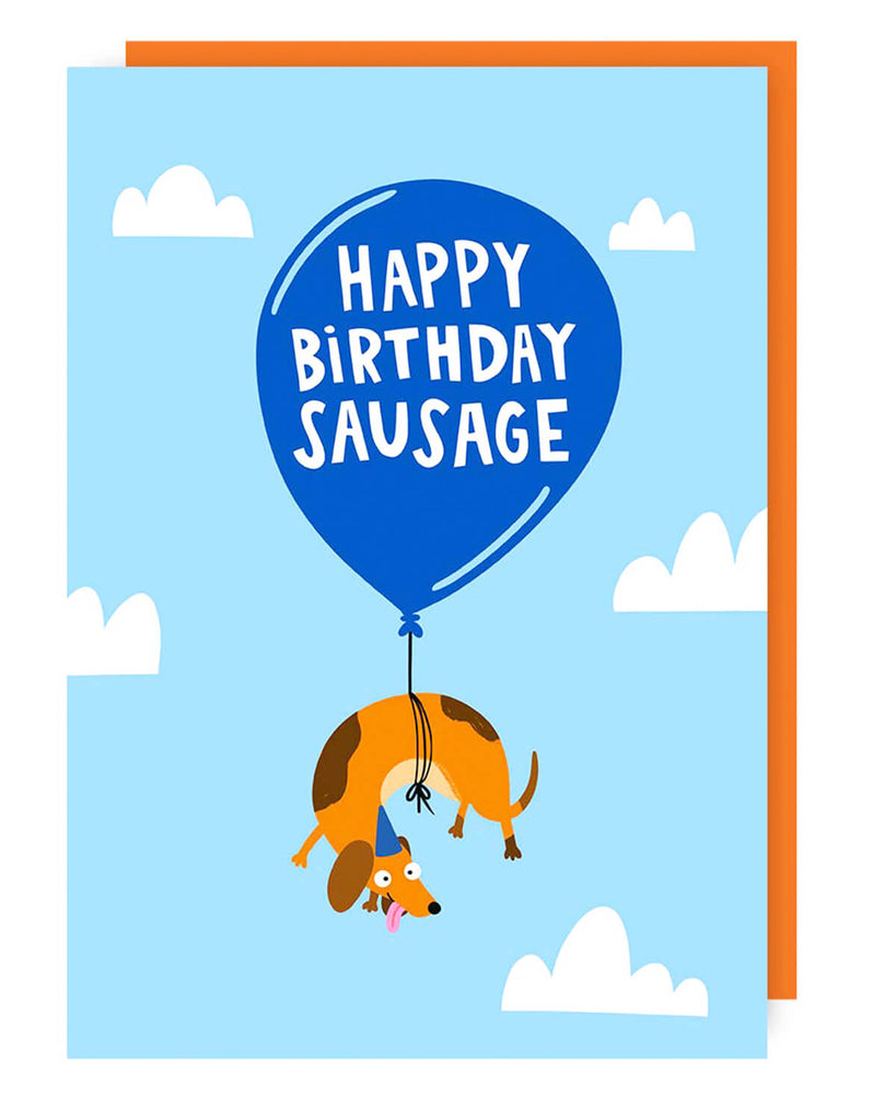 A fun birthday card featuring a cartoon sausage dog wearing a party hat, floating in the sky while hanging from a blue balloon with “HAPPY BIRTHDAY SAUSAGE” written on it. White clouds decorate the bright blue background, and an orange envelope is included.
