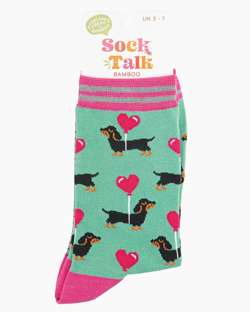 Packaged mint green bamboo socks featuring black and tan sausage dogs carrying pink heart balloons, displayed with a “Sock Talk” label.