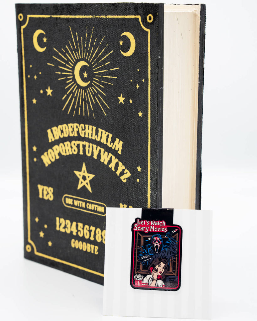 Let’s Watch Scary Movies magnetic bookmark from Rebel Romance shown in front of a gothic-style book with a Ouija board design. The bookmark features a horror-inspired comic illustration of a woman on the phone with popcorn, while a masked killer lurks behind her. A spooky and fun accessory for horror fans and readers who love a good scare. Perfect for alternative millennials and Halloween vibes all year round.