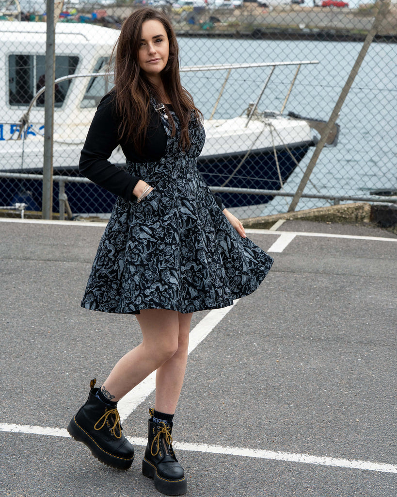 Amy stands outdoors near a harbour wearing a black flared pinafore with a grey mystical creature print, paired with chunky black boots and a long sleeve black top. She poses with one hand in her pocket while the skirt flares gently.