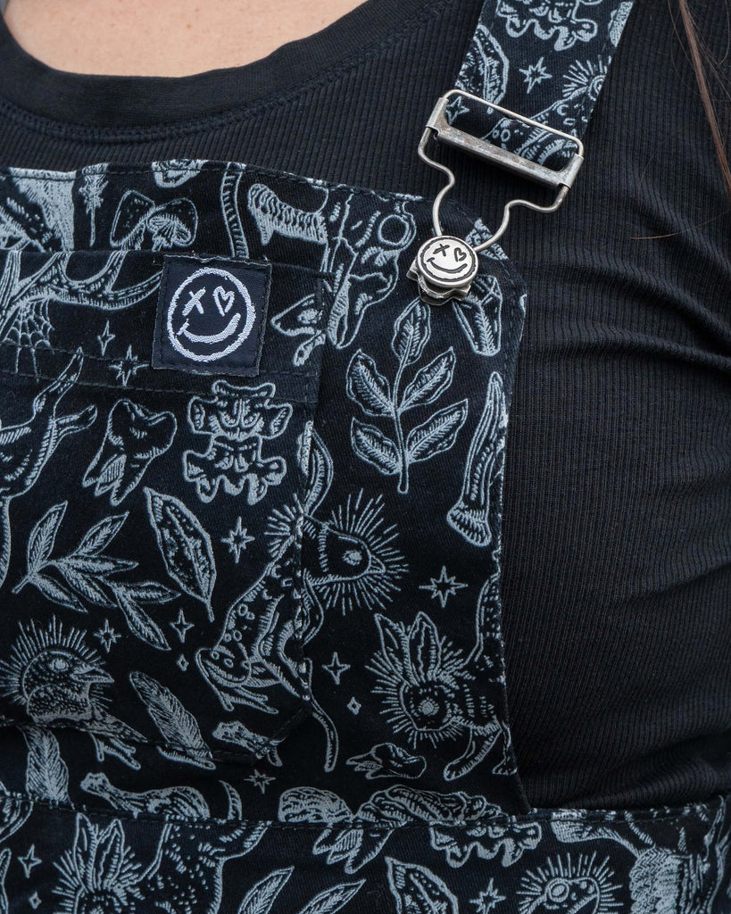 Close up of the Seek pinafore bib showing the grey illustrated print of birds, mushrooms and magical motifs on black fabric. The metal strap buckle with a smiley face button is also visible against Amy’s black top.