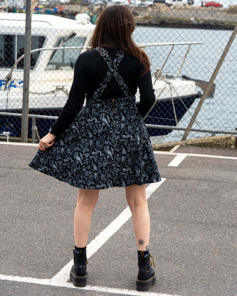 Rear view of Amy wearing the Seek flared pinafore with criss cross straps and a skater style skirt. The mystical grey print covers the whole pinafore as she stands near boats and fencing at the harbour.
