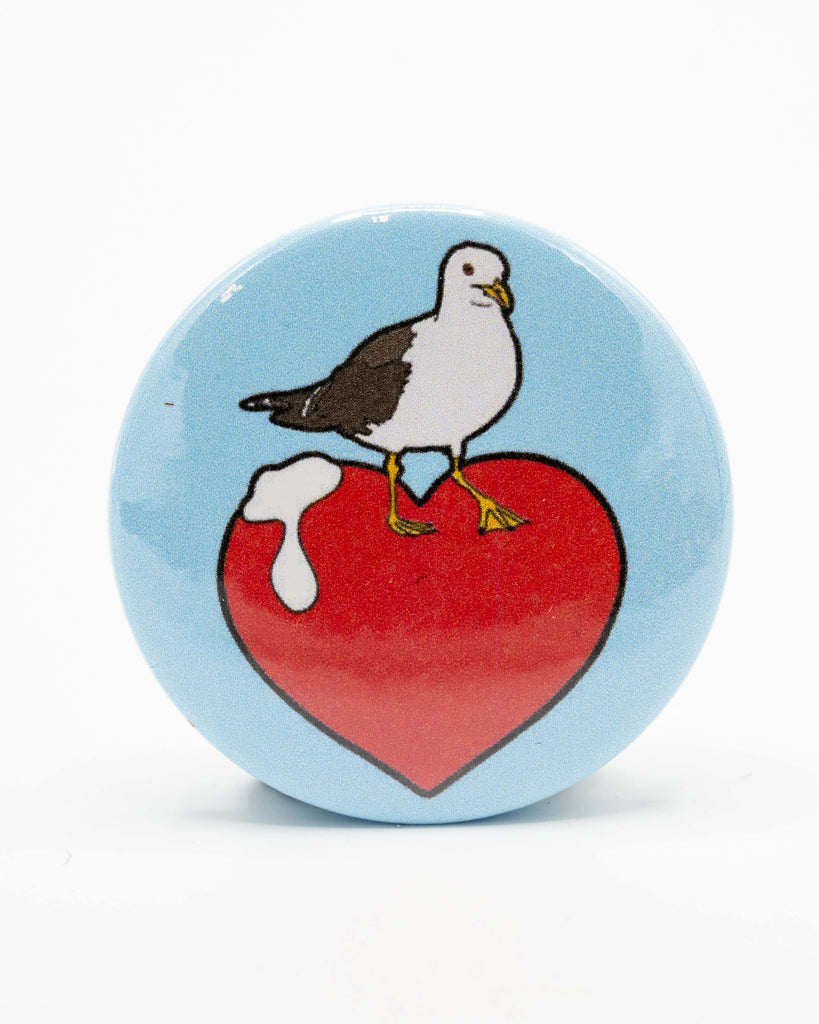 Rebel Romance "I Love Seagulls" button pin badge by Bite Your Granny, featuring a seagull standing proudly on a red heart with a splat of seagull poop. The badge has a blue background and bold cartoon-style illustration. Perfect for fans of British seaside humour, this playful pin adds a quirky touch to any jacket or bag. From a UK brand that champions extraordinary and alternative style.