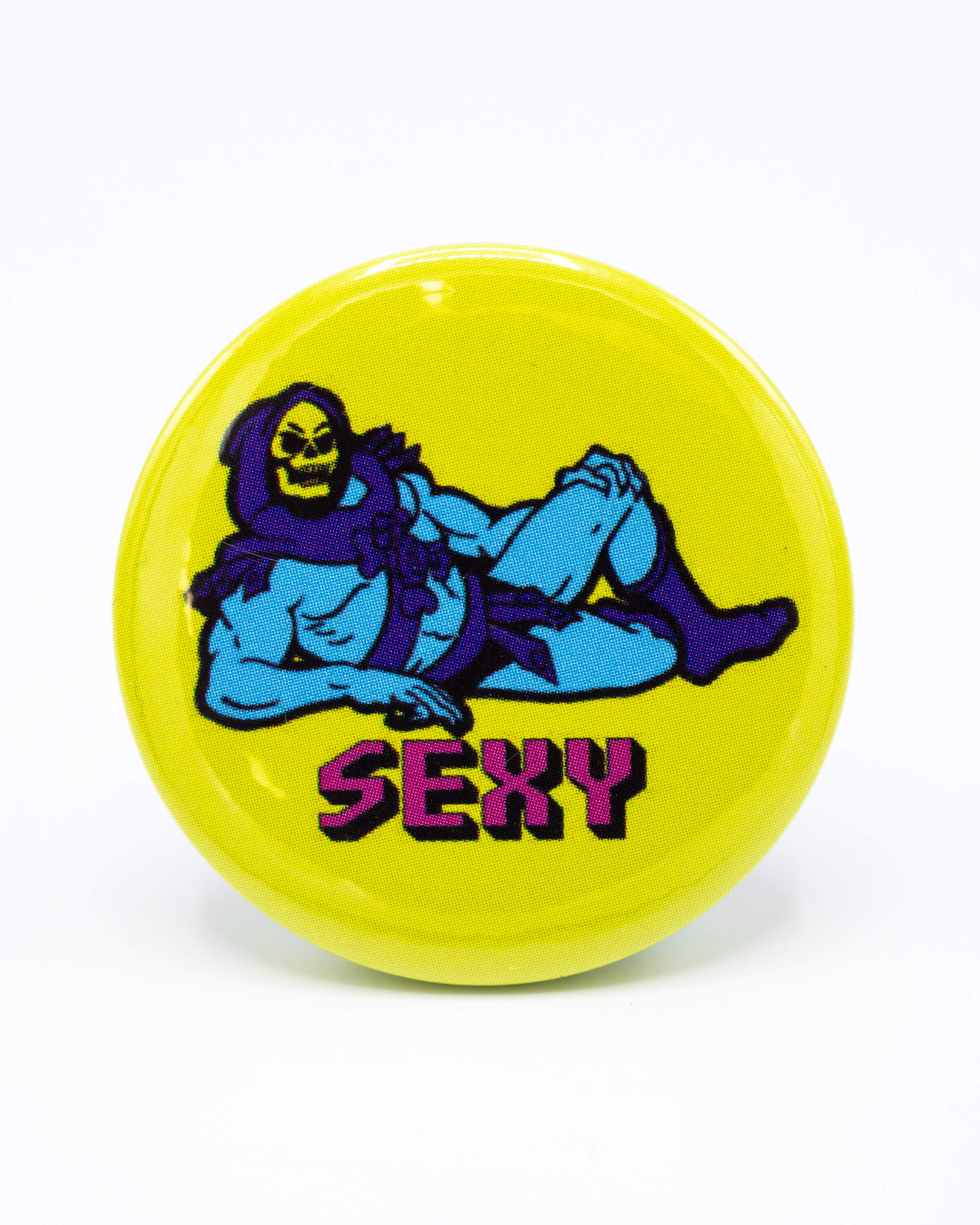 Yellow button badge with a skeletor cartoon character laying down and 'SEXY' pink text underneath. An 80s inspired retro design from Bite Your Granny.