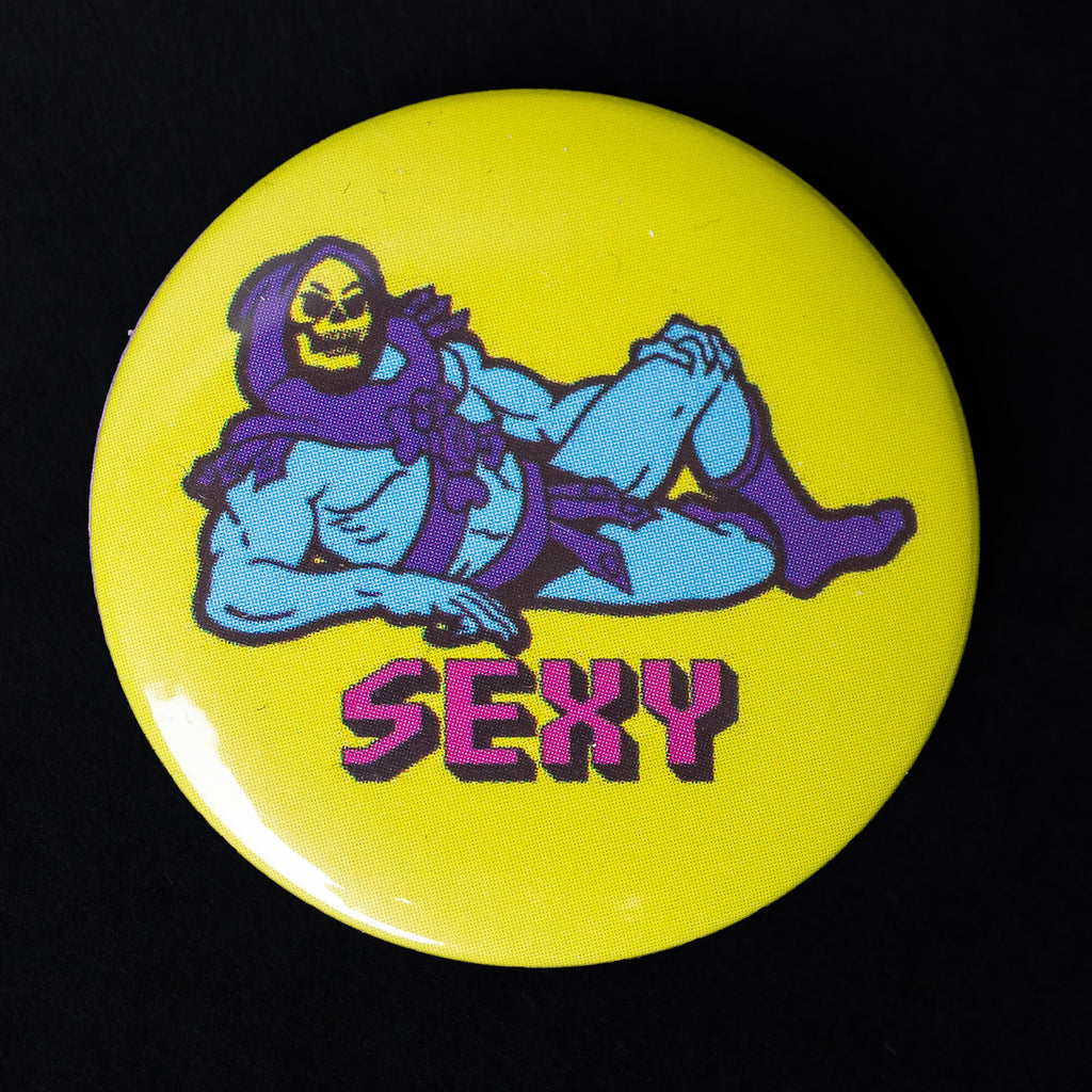 A 38mm button badge featuring Skeletor lounging provocatively on a bright yellow background with the word “SEXY” written in bold pink retro-style letters. This high-gloss pin is a cheeky nod to pop culture, perfect for personalising your accessories. Designed by Bite Your Granny.