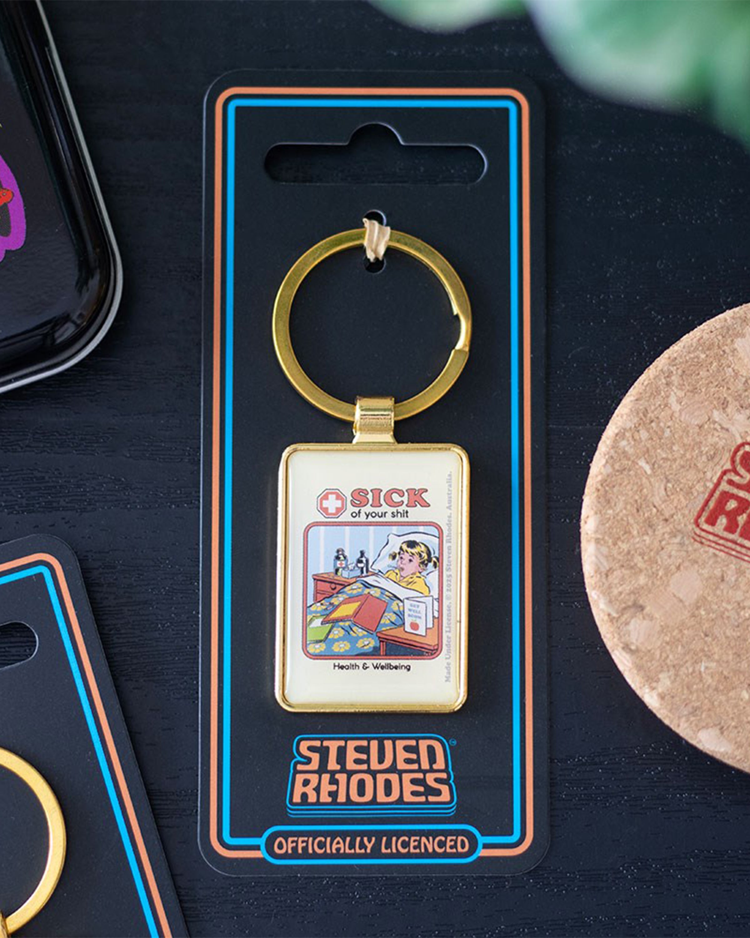 Steven Rhodes “Sick of your shit” enamel keyring shown in packaging on a dark table alongside other accessories. Retro-style humour gift, officially licensed and available at Rebel Romance, a UK alternative brand selling fashion and quirky homeware.