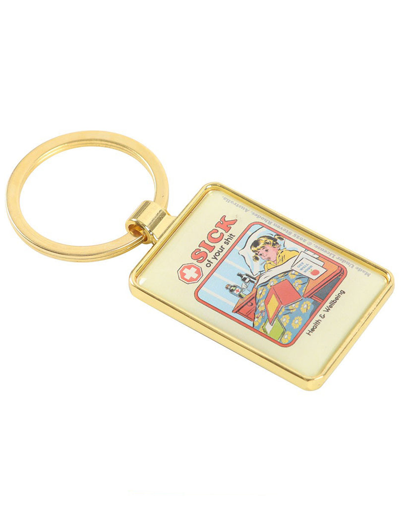 Angled view of the Steven Rhodes “Sick of your shit” enamel keyring with gold edging. Retro comic art shows a sick child in bed, surrounded by medicine and books. Available at Rebel Romance, a UK alternative lifestyle store for millennials.
