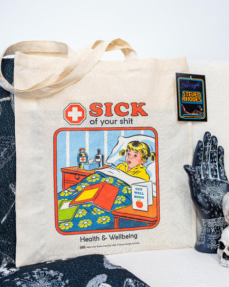 A natural cotton tote bag featuring Steven Rhodes’ retro-style artwork. The design shows a child in bed with a “Get Well Soon” card and the bold text “Sick of Your Shit” above, with “Health & Wellbeing” written below. The bag is eco-friendly and officially licensed merchandise.