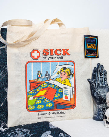 A natural cotton tote bag featuring Steven Rhodes’ retro-style artwork. The design shows a child in bed with a “Get Well Soon” card and the bold text “Sick of Your Shit” above, with “Health & Wellbeing” written below. The bag is eco-friendly and officially licensed merchandise.