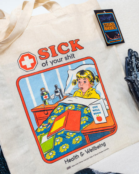 A natural cotton tote bag featuring Steven Rhodes’ retro-style artwork. The design shows a child in bed with a “Get Well Soon” card and the bold text “Sick of Your Shit” above, with “Health & Wellbeing” written below. The bag is eco-friendly and officially licensed merchandise.
